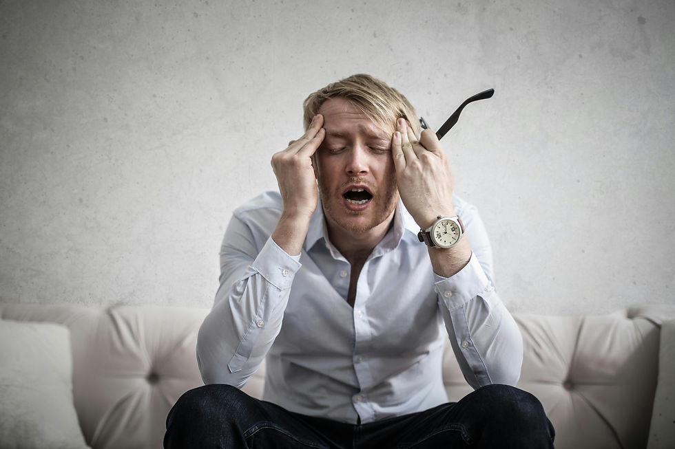 Autistic man in white shirt sitting on beige couch, holding head in frustration. Background is a plain wall. Emotional expression of stress.