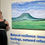 Thumbnail: Image shows Kerry Valley on display mounted on the wall with the text "Natural Resilience: inner feelings, outward reflections. Chloe Wigg" on the left hand side of the image is artist Chloe Wigg in a black cocktail dress and white belt holding a glass.