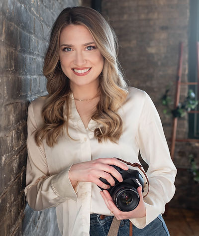Woman standing next to brick wall in white blouse holding a camera