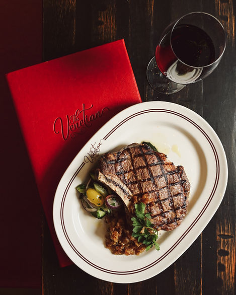 Steak and vegetables on a white plate sitting on a red menu book with a glass of red wine