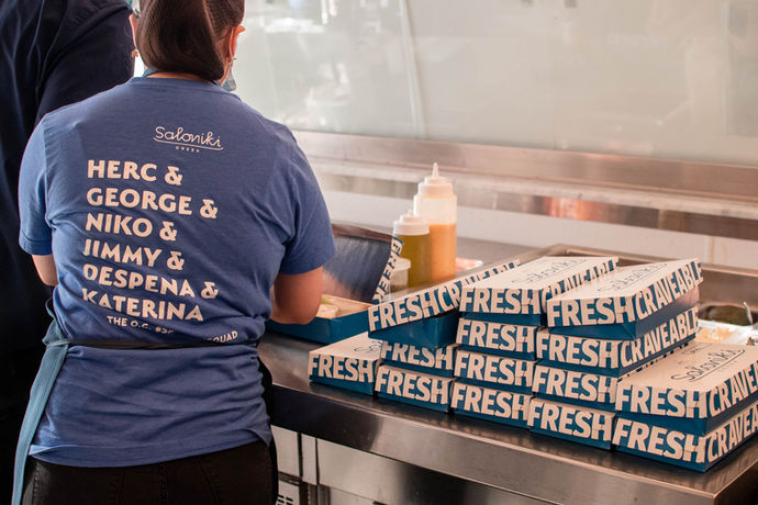 Employee in a branded blue t-shirt plates dishes into blue takeout containers that says "fresh" on the outside