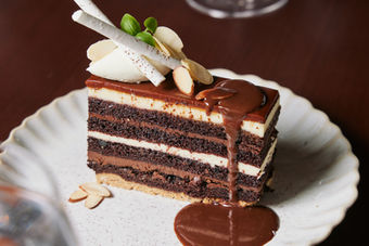 Chocolate cake slice with chocolate sauce and toppings on white plate.