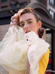 A model poses with sheer white fabric billowing around them, wearing a bold yellow outfit and vibrant yellow-green eye makeup. Their gaze is intense, with anuran architectural backdrop adding depth to the image. 