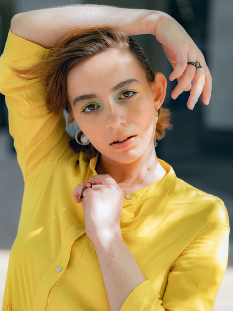 A model wearing a yellow outfit poses with one arm raised above their head, gazing directly at the camera. Their bold yellow-green eye makeup contrasts beautifully with their natural complexion, set against a blurred urban background. 