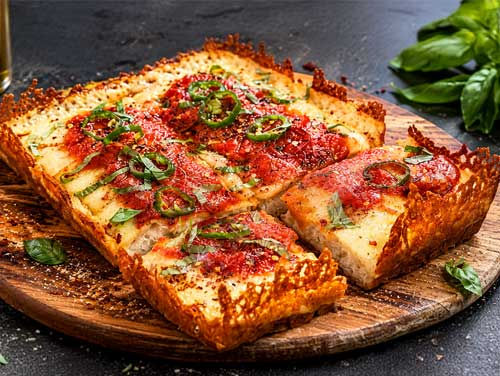 Detroit-style pizza with crispy cheese crust, tomato sauce, basil and green chilli on a wooden board, with chili flakes and basil leaves nearby.