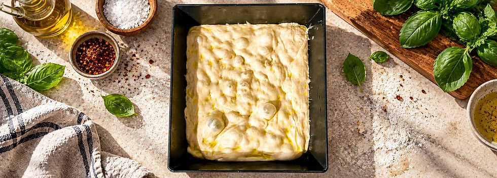 Fresh focaccia style dough in a Detroit pan on a sunlit countertop, surrounded by basil, olive oil, salt, and chili flakes