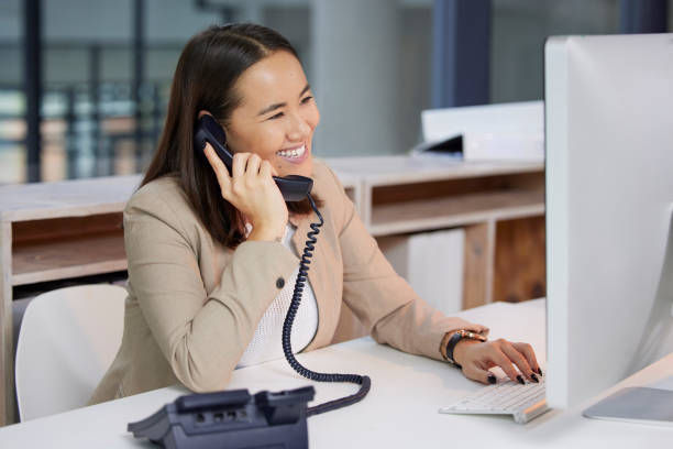 Smiling woman in a beige blazer talks on the phone and types on a computer in a modern office with a bright, open background.