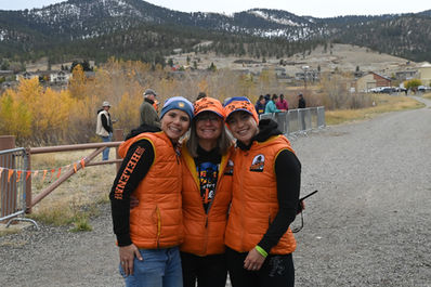 Three smiling women in orange vests, outdoors with mountains.