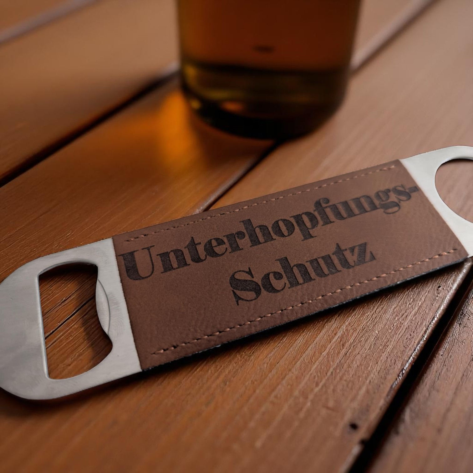 Gravierter Edelstahl Flaschenöffner mit Lederapplikation und Schriftzug „Unterhopfungs-Schutz“ auf Holztisch