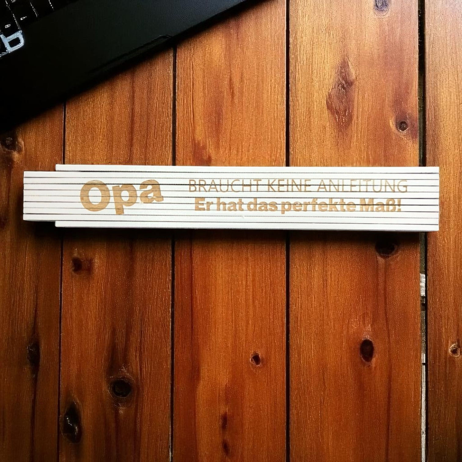 Weißer gravierter Holz-Zollstock mit Spruch für Opa liegt auf Holzwand, Lasergravur „Opa braucht keine Anleitung – Er hat das