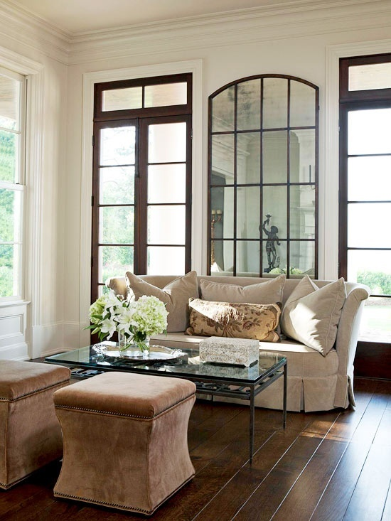 Elegant living room with a beige sofa, floral pillow, glass coffee table, flowers, and arched windows. Warm, cozy atmosphere.