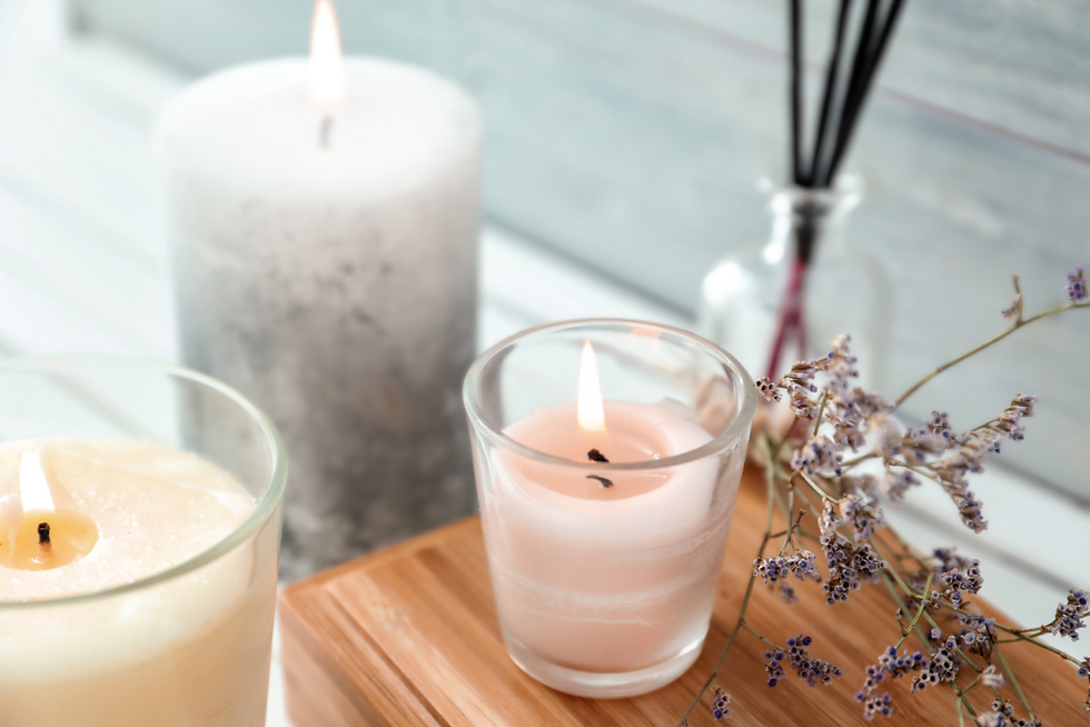 Lit candles in glass holders on a wooden tray with dry lavender sprigs. Background shows a muted gray wall, creating a calm ambiance.