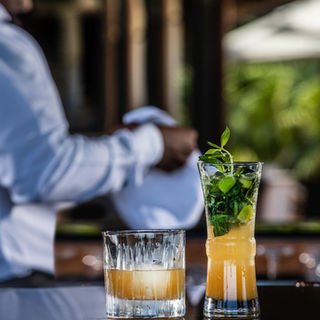 Image of a bar, with 2 rum cocktails displayed proudly. In the background is the bartender, holding a cloth.