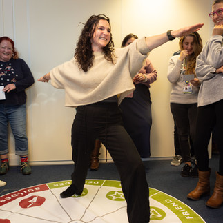A workshop participant actively engaging with a large floor mat depicting the Motivational Maps clusters. She is striking a fun pose while balancing on a section of the map, while colleagues laugh and support her. This illustrates an interactive, kinetic learning exercise designed to help teams understand their motivational drivers.