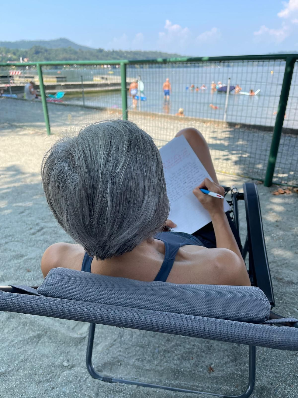 Susannah Brade-Waring, author of The Confident Manager, in a lounge chair writes in a notebook by a lake, with swimmers and green hills in the sunny background.