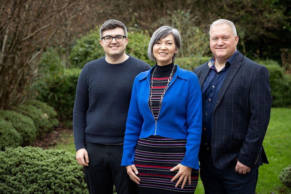 Three smiling adults stand together in a garden, with shrubs and trees behind them; one wears a bright blue jacket.