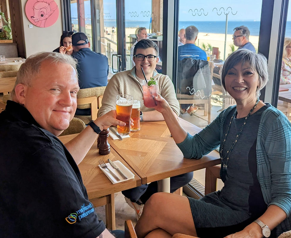 Three friends smile and toast with beer and a pink cocktail at a seaside restaurant with ocean view.