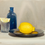 Thumbnail: a still life oil painting depicting a blue bottle a lemon on a silver plate and a glass with a slice of lemon balanced on top
