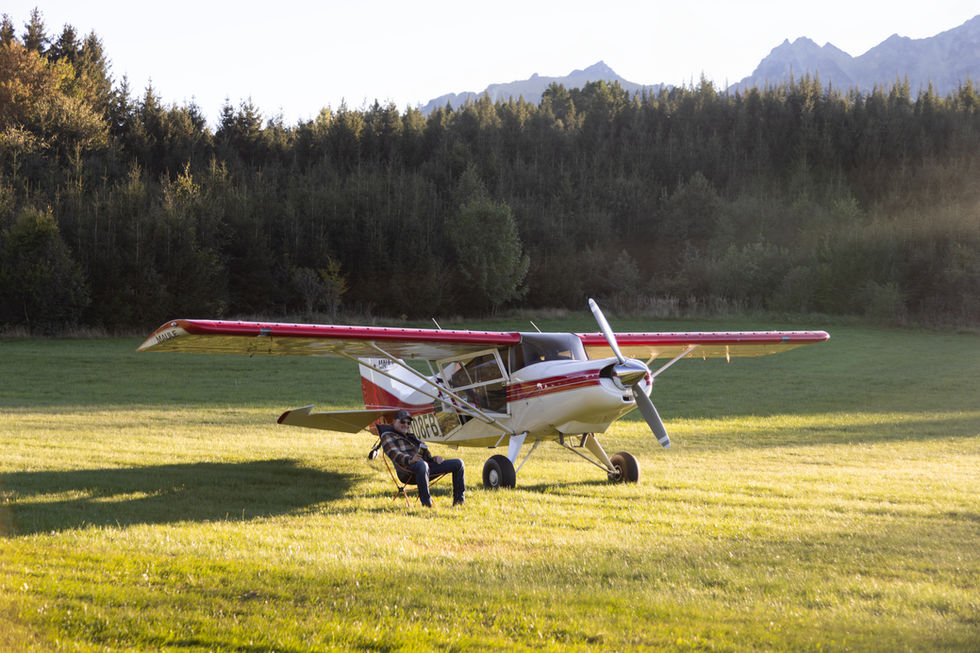 Maule MX7 aircraft, registration N108FB side view, parked on a grassy field at sunset, pilot sitting in a chair next to plane, photo by SamyKramer