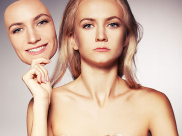 A woman with a serious expression holds up a smiling face mask beside her own face, symbolizing the hidden emotional cost of masking.