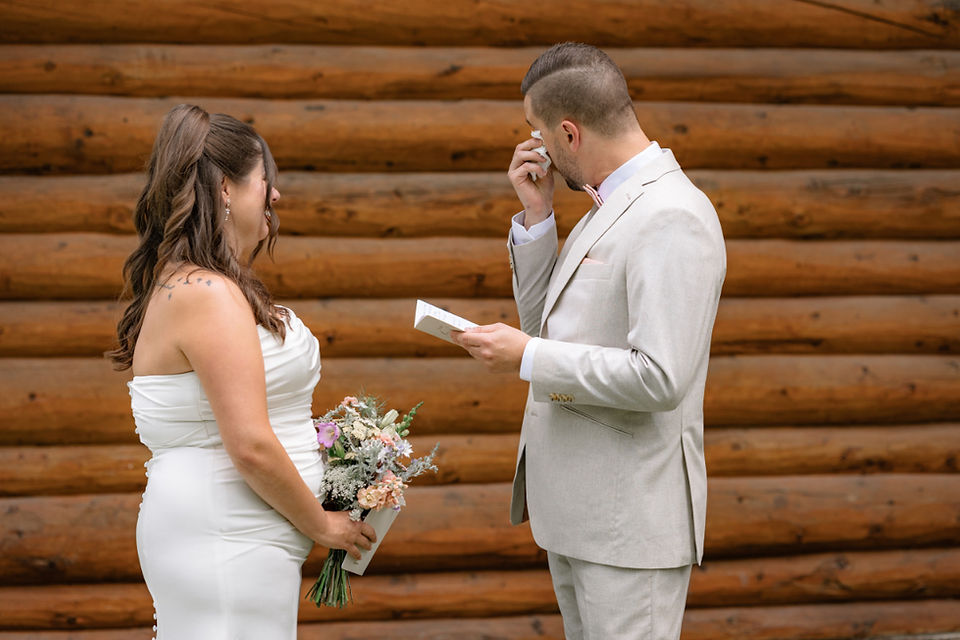 Groom wiping away tears while reading vows to bride in front of rustic wooden cabin