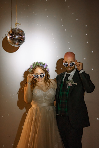 Newly married couple wearing playful sunglasses and dancing under disco ball lights at a fun and intimate wedding reception