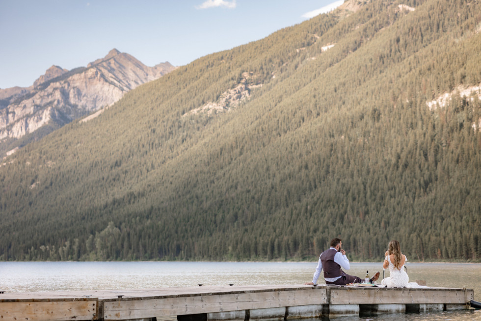 Wide lake view of a couple sitting on a dock during eloping in Banff, celebrating with a quiet mountain lakeside picnic.