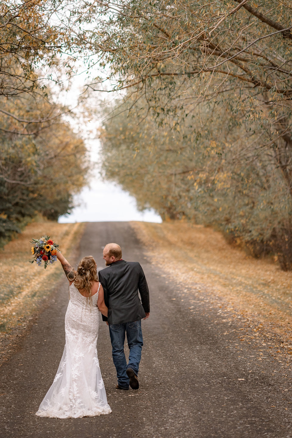 Bride and groom walking hand in hand down a tree lined road, photographed by a destination wedding photographer during golden hour.