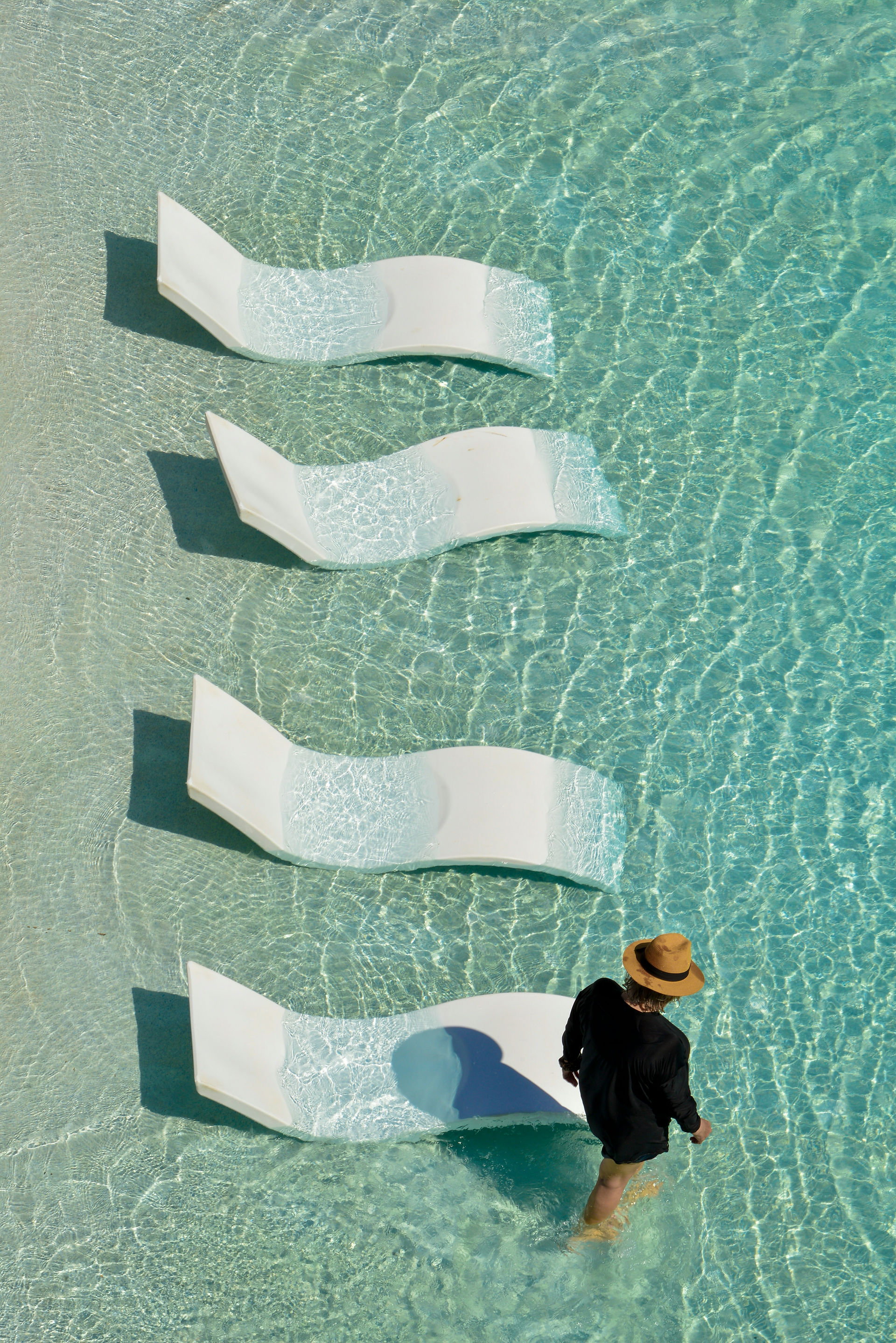 Overhead view of a woman in a hat walking beside white sun loungers submerged in a turquoise swimming pool.