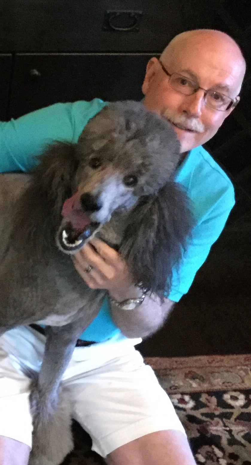 An image of David Henson, a person with glasses and a white mustache smiling into the camera. He is accompanied by a dark gray poodle.