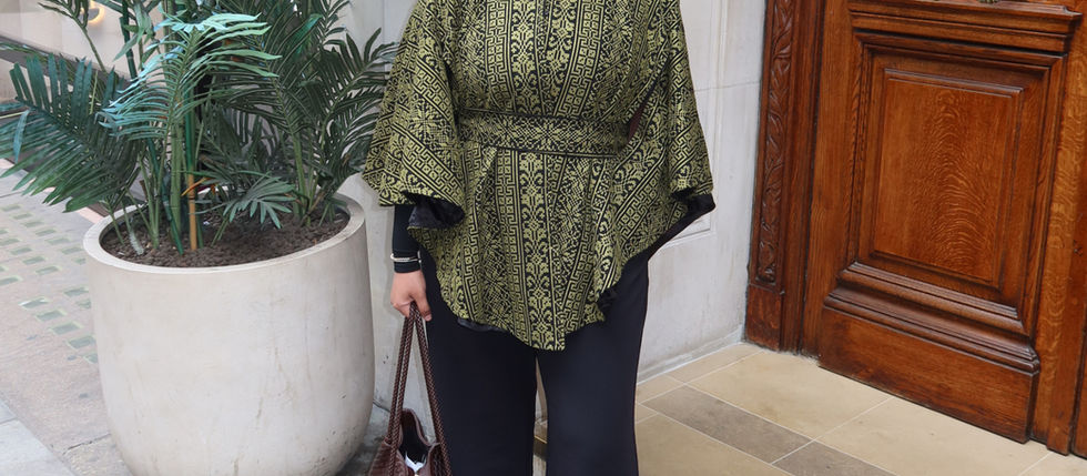 Shereen Patel stands outside a building entrance in London, wearing a patterned green and black top, black trousers and a brown headscarf, carrying a woven brown handbag. Tropical plants sit beside her as she looks towards the camera.