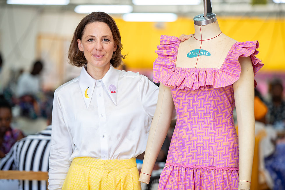 Joanna Maiden stands beside a mannequin wearing a pink patterned dress inside the SOKO Kenya factory, smiling with her hand resting on the mannequin’s shoulder, with workers and machinery visible in the background.