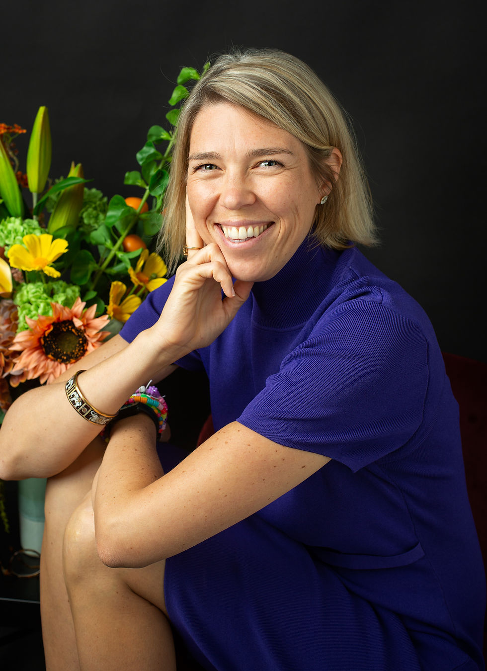 Close-up portrait of Elisabeth Prager, founder of Mad Money Club, smiling towards the camera in a purple dress, seated beside a colourful floral arrangement against a black background.