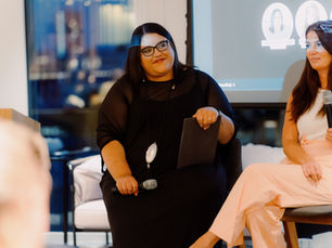 Tara Attfield-Tomes sits on a panel in a black outfit, holding a microphone and a black folder. She wears glasses and a statement necklace, and is smiling thoughtfully. Beside her is another panel speaker. Behind them, a screen displays a speaker lineup with Tara’s name visible. The image captures Tara’s presence as a confident leader and changemaker in the funding and entrepreneurship ecosystem.