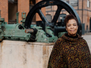 Dr Marilyn Comrie standing outdoors beside industrial machinery, with a large mechanical wheel and brick buildings in the background.