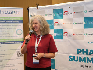 Suzie Harvey speaking on stage at a life sciences conference, holding a microphone and presenting to an audience. She is standing in front of event branding and sponsor displays, wearing a red top and conference lanyard.