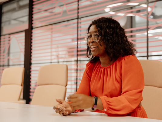 Lisa Maynard-Atem sitting at a table in a modern meeting room, mid-conversation, wearing a red dress and glasses, gesturing as she speaks.