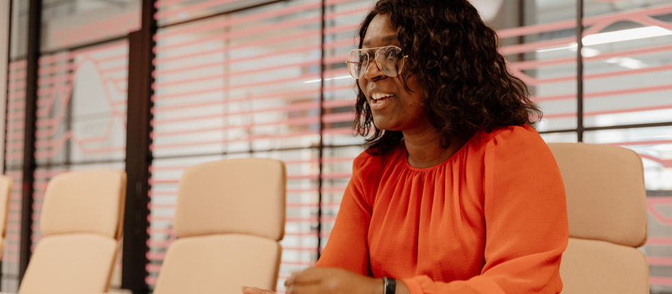 Lisa Maynard-Atem sitting at a table in a modern meeting room, mid-conversation, wearing a red dress and glasses, gesturing as she speaks.