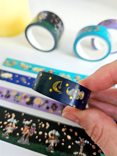 Hand holding washi tape which is dark blue with moon, stars and clouds on tape. Other washi tapes of various colours shown unfocused in background.
