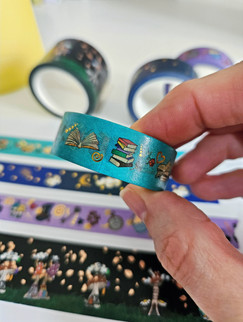 Hand holding a light blue washi tape with a design on it of various book illustrations. Other washi tapes of various colours shown unfocused in background.