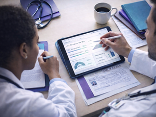 Doctors in white coats review data on a tablet with charts and graphs. Papers, a stethoscope, and coffee cup lie on the desk.