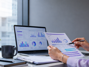 Person reviews colorful graphs on paper and laptop in an office. A plant, mug, and notebook are nearby on the desk. Bright and focused mood.