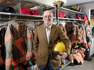 A white man in his 70's with glasses and a bald head, smiling at the camera with a yellow firefighter helmet in his hand standing in front of fire fighter bunker gear.