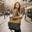 Thumbnail: Woman walking on a street, holding a small canvas bag, with buildings in background.