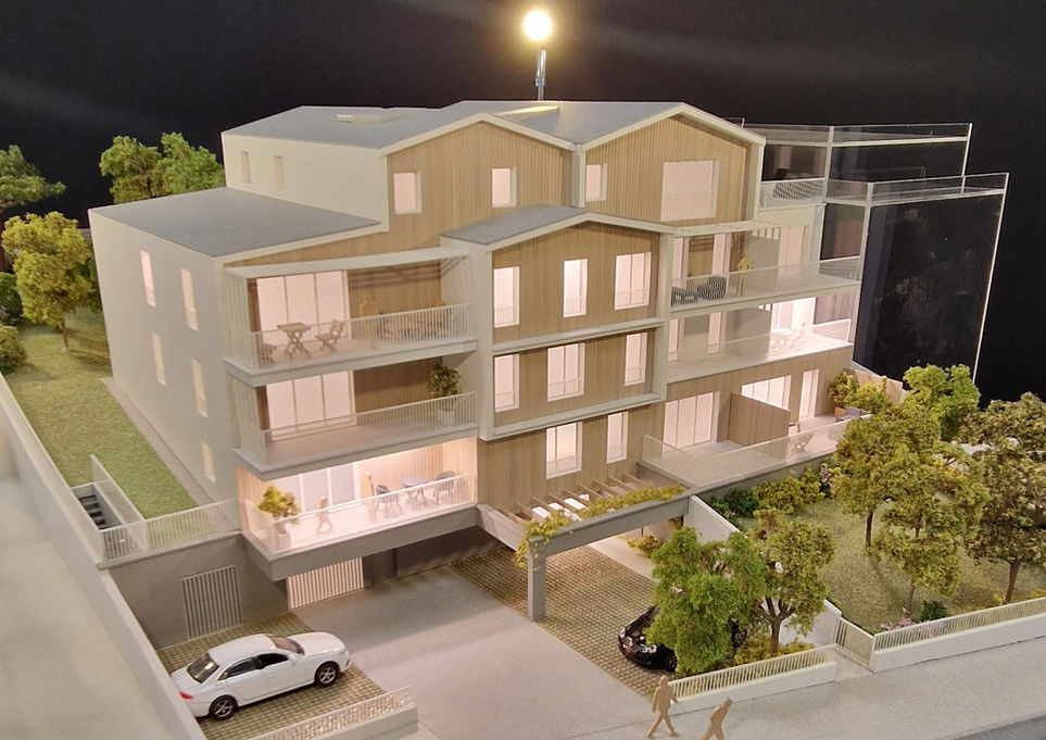 Vue de la maquette illuminée du projet "L'Initial" à Saint-Marc-sur-Mer : résidence intimiste à l'architecture balnéaire moderne mariant bois et façades blanches, dotée de larges balcons et d'un jardin arboré en bordure d'avenue.