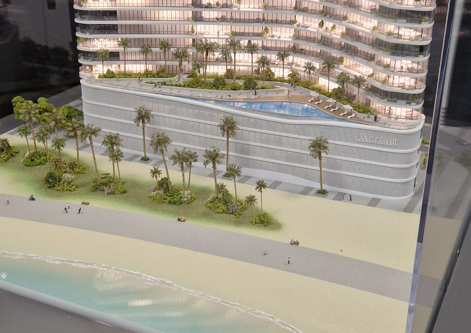 Maquette illuminée sous vitrine de la tour résidentielle de luxe "Marlin 2" à Abu Dhabi : architecture aux lignes ondulées évoquant les vagues, surplombant une piscine à débordement sur podium et une plage de sable fin bordée de palmiers et d'eau turquoise.