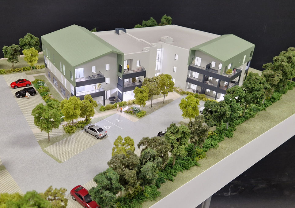 Vue de la maquette illuminée de la résidence intimiste "Hélia" à Laval : petit immeuble collectif aux toitures vertes et balcons foncés, intégré dans un environnement dense et arboré avec parking paysager.