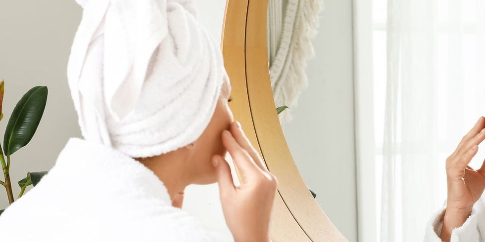 Woman in a towel and robe looking at her skin in the mirror during a self-care routine.