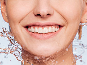 Woman with radiant skin smiling as water splashes around her face, representing deep hydration and natural glow after skin booster treatments at Luminous Skin in Nelson.