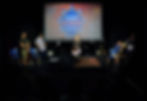 an image of a woman speaking a dark stage, with a bright light on her